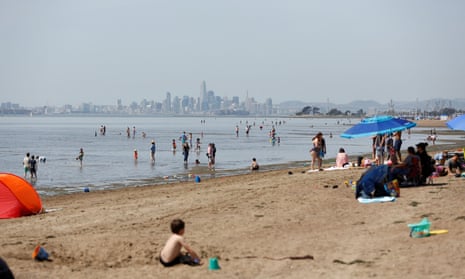 People sit on a beach and enjoy wading in the water. In the distance, the San Francisco skyline can be seen.