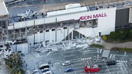 Rescue workers gather on a debris-covered street beside a severely damaged white building with collapsed walls