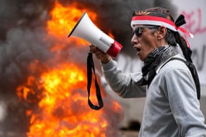 A man shouts slogans into a megaphone with flames burning in the background