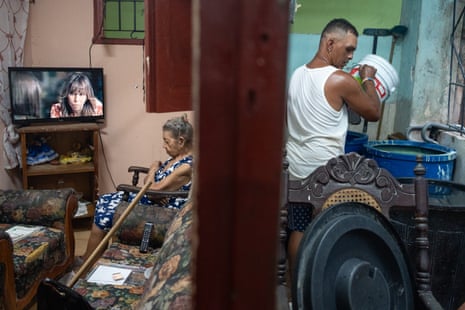 A man in a white vest top pours water into a blue drum in a small flat in Havana, while an elderly woman watches TV in the room next door.