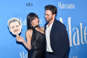 Dakota Johnson holds a cutout of Pedro Pascal’s head as she embraces actor Chris Evans by a blue wall
