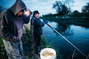 Dusk by a river. A man uses a torch to look at a bucket of elvers as another man handles a net on a long pole