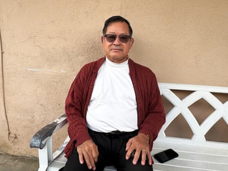 Omar Muñoz, wearing sunglasses and a maroon cardigan, sits on a white bench outdoors.