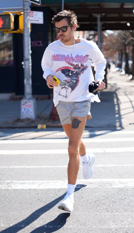 Harry Styles in sunglasses jogging over a road crossing wearing a white sweatshirt with graphic print, grey shorts and white trainers and socks while holding a phone and with headphones on