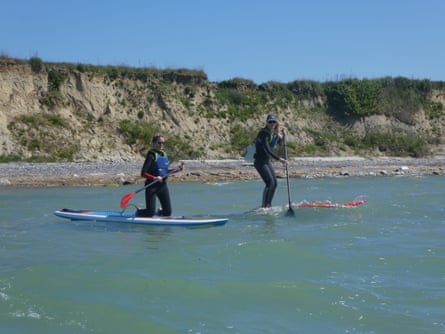 One person kneeling on a paddleboard and another standing, close to a shore with a low, chalky cliff
