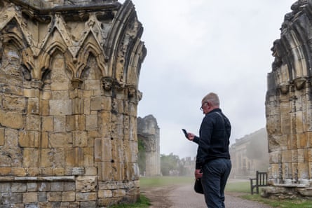 A man stands looking at his phone in Museum Gardens, York, between stone pillars and remains of historic buildings; it is misty in the background and the scene appears atmospheric.