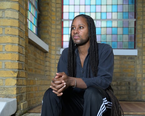 Jason Arday sits on a step in front of a window with small blue, yellow, green, pink and gold panes of stained glass and by a pale yellow brick wall. He has very long braids and wears a black shirt and tracksuit trousers with white stripes down the side. He has his hands clasped over one knee and looks thoughtful.