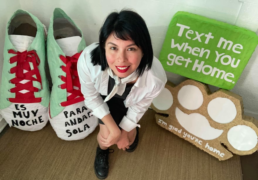 Benavidez sits between artworks including a huge pair of shoes that say 'es muy noche' on one shoe's toe and 'para andar sola' on the other. on her other side is a sign that says 'text me when you get home' and a design that looks like a pawprint that says 'i'm glad you're home' under it