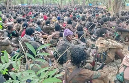 Armed men in combat clothes sit on the forest floor at a meeting