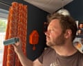 A man holds a small fan up to his face while standing near orange-patterned curtains in a dark-walled room