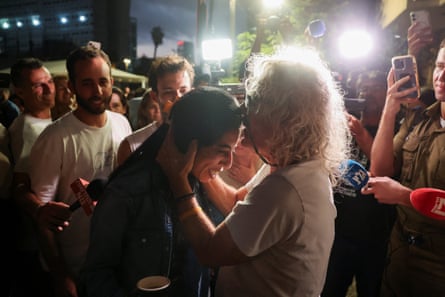 One person kisses another on the forehead in front of a group of people watching under camera spotlights