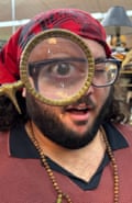 A man with a beard, glasses and a red T-shirt on his head putting a magnifying glass to his eye.