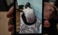 A hand holds a photograph up to the camera of a severely emaciated man sitting with his back to the camera on a bed