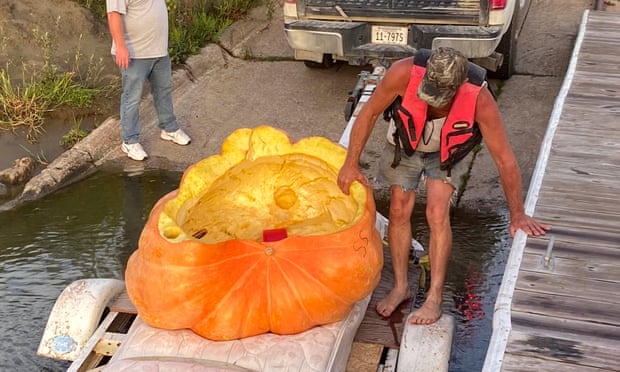 man in lifejacket holds on to giant pumpkin near dock