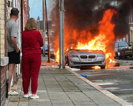 A man and a woman stand by the front door of a house on McMaster Street and watch as a car burns.