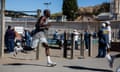 a tall black man in a tank top shorts and sneakers runs on a track with fencing and people behind him