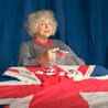 Portrait of Miriam Margolyes holding a broken union flag teacup while sitting at a table with a union flag tablecloth