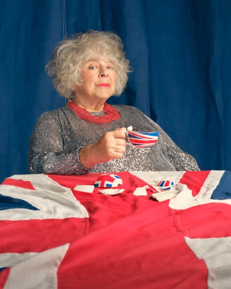 Portrait of Miriam Margolyes holding a broken union flag teacup while sitting at a table with a union flag tablecloth