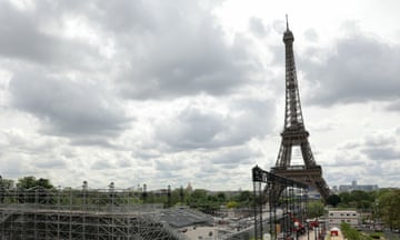 The Eiffel Tower is seen silhouetted against a grey, cloudy sky with construction work under way in the foreground; long sections of scaffolding and temporary huts and structures are seen.