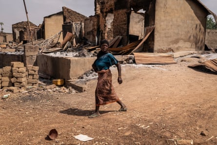 A woman in a blue shirt and brown skirt walks past debris, burnt structures and damaged shops.