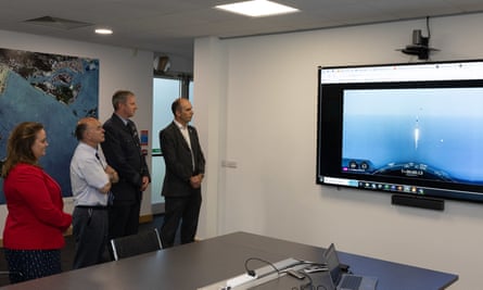Four people watching a screen on a wall showing a distant view of a rocket launch over the sea