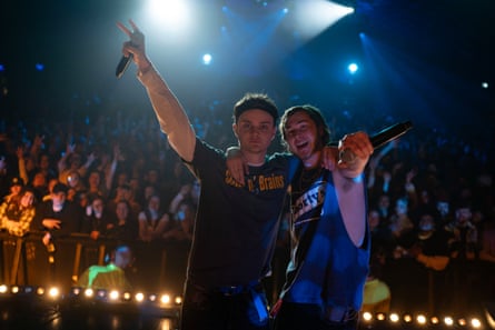 Standing on stage in front of a large crowd, they embrace, Bottomley pointing at the camera, Ross gesticulating into the air