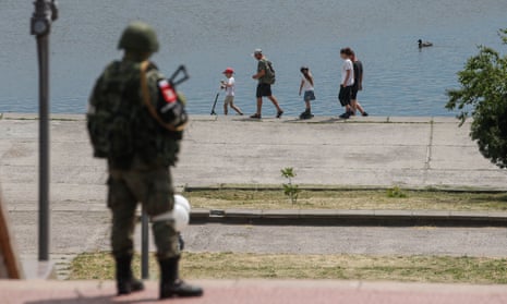An armed Russian soldier in combat gear watches a family strolling along the Dnipro River in Kherson.