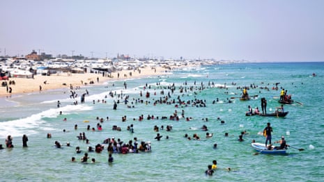 A large number of people in the sea, some of them in boats, and behind the a beach lined with displaced people’s tents