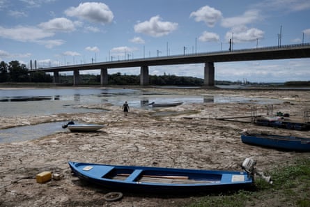 A man walks along the dry river bed behind a small blue boat, embedded in algae and mud. A road bridge spans the dried-up river which has been reduced to shallow puddles and a small stream of water.