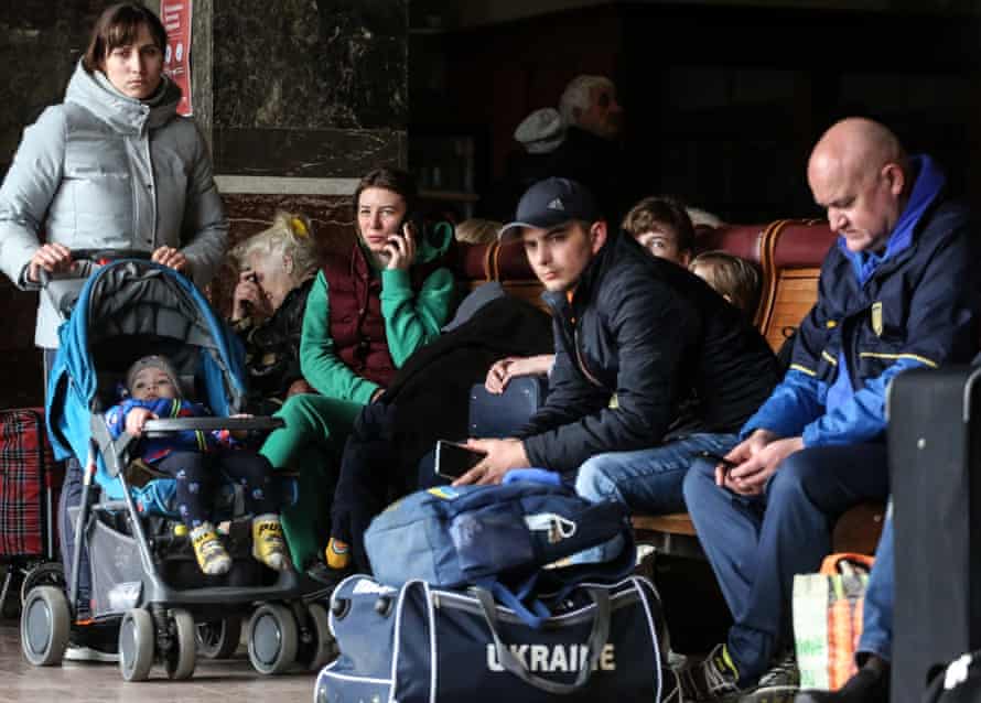 Three women, one standing with baby in buggy, and two men sitting in the station waiting room.