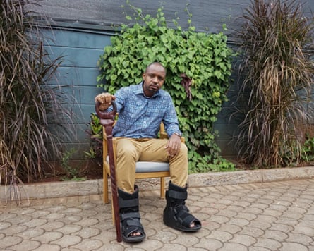 Boniface Mwangi sits on a chair holding a walking stick and wearing foot braces
