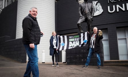 Two supporters with flag saying ‘You’ll never ever beat Dan Burn’ beneath statue with another man in foreground outside Newcastle ground