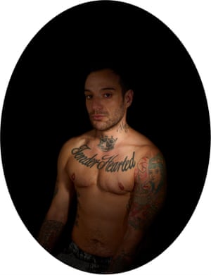 Transgender man Rocco Kayiatos sitting in front of a black backdrop and looking at the camera as light falls on him and his tattoos, which included the words 'Tender Hearted' across his chest