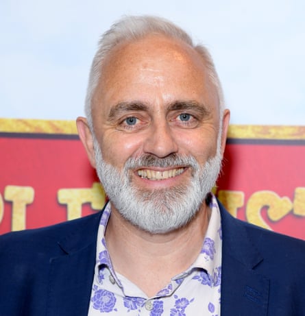 A man with grey hair and a grey bear smiling, wearing a dark blue jacket and a white shirt with a blue pattern