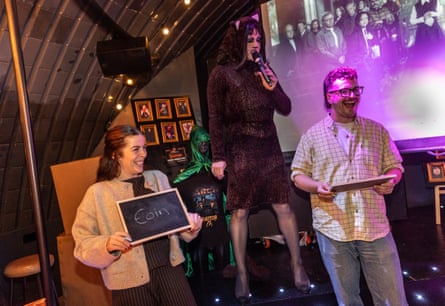 A host in a dress and cat makeup stands between two smiling contestants, each holding a chalkboard with a name on it