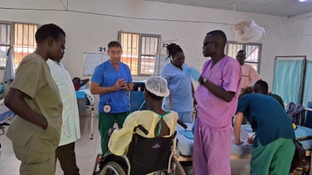 A doctor in blue scrubs speaking to a patient in a wheelchair facing away from the camera