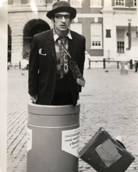 A black and white image of a man in a crumpled hat and suit emerging from a waste bin with a briefcase propped against it.