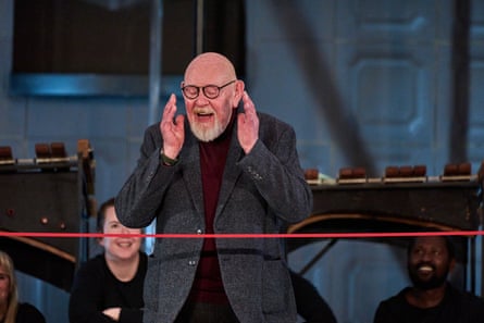 Mark Dornford-May claps with his hands held high to his face as he awaits the cutting of the red ribbon. He has a white beard and wears glasses.