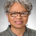 A headshot person with short gray hair wearing glasses and earrings