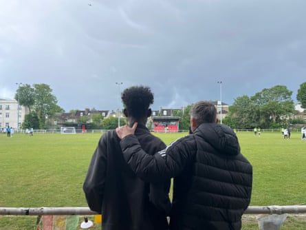 They stand behind a pole surround a football pitch, looking at players on the pitch. The man on the right has his arm around the shoulder of the man on the left