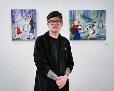 A young man dressed in black, with glasses and tattooed forearms, stands in front of two pictures at a gallery