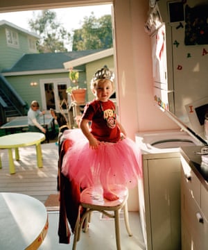 A young boy wearing a pink tutu and a silver crown stands on a chair beside a washing machine, looking at the camera and smiling