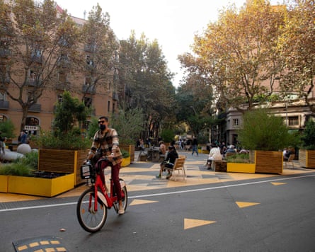 A man rides a bicycle in a pedestrianised part of Barcelona marked by yellow tree planters and road markings in a bright tree-lined intersection