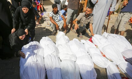 Seven bodies wrapped in shroud, with a woman crouched over one and crying as she is held by another woman
