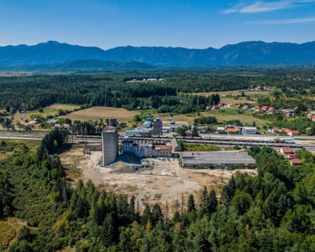 A green landscape with an industrial estate in the foreground, surrounded by green trees and mountains visible in the background.