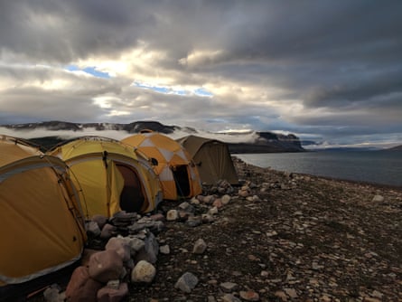 A line of four dome tents on a rocky beach in low light in a barren Arctic landscape.
