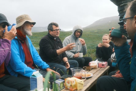 A group of men sat at a small wooden table outside listening to a priest talking A group of men sat at a small wooden table outside listening to a priest talking