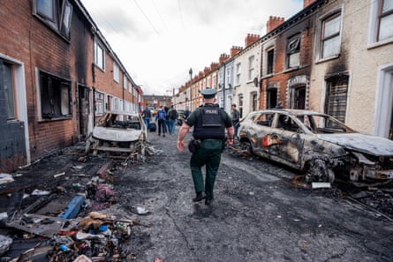 A police officer walks away from the camera down a residential street with burnt-out cars and homes