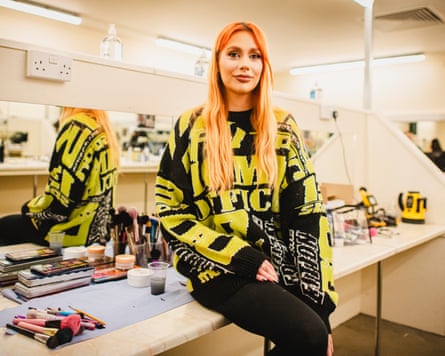 A woman with ginger hair and a neon green and black jumper sits on a desk surrounded by makeup.