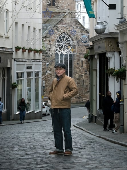 A man wearing a newsboy cap, jeans and a light brown jacket stands in the middle of a cobbled street
